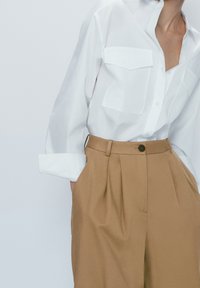 Vit knapp-up skjorta med två bröstfickor, matchad med högmidjade beiga byxor med veck och en ensam brun knapp.