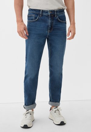 Homme portant un jean bleu foncé retroussé et des baskets blanches, debout devant un fond clair uni.