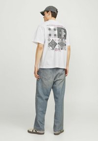 Camiseta blanca de algodón con estampado gráfico colorido en la parte posterior, que presenta formas geométricas y texto; combinada con unos vaqueros de mezclilla azul holgados.