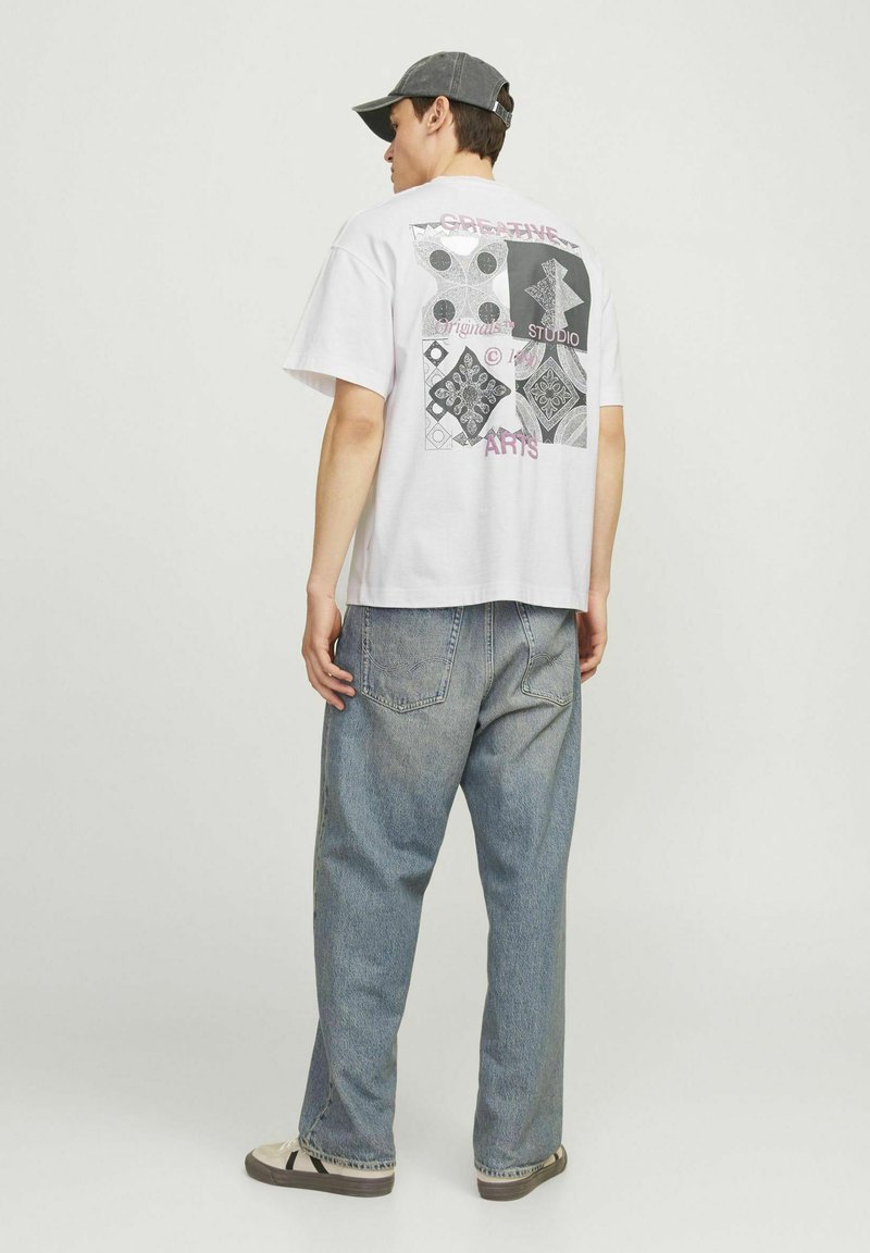 Camiseta blanca de algodón con estampado gráfico colorido en la parte posterior, que presenta formas geométricas y texto; combinada con unos vaqueros de mezclilla azul holgados.
