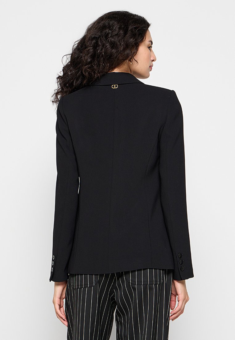 Veste noire sur mesure avec un col cranté, design ajusté, et un accent doré à l'arrière. Comprend des manches longues et des poignets à boutons.
