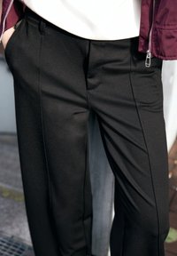 Pantalon noir sur mesure en tissu texturé, avec deux poches latérales, de subtils plis frontaux et une coupe ajustée.
