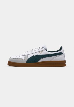 Fehér sneaker zöld velúr részletekkel és perforációkkal. Barna gumi talppal és fűzős záródással rendelkezik. Simább textúra kontrasztos részletekkel.