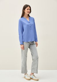 Camicia a maniche lunghe azzurro chiaro con colletto henley e dettaglio dei bottoni, abbinata a jeans grigi chiari a gamba larga e sneakers in tonalità neutre.