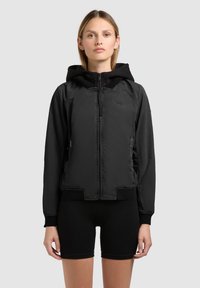 Ung kvinde iført en sort hættetrøje med lynlås og hætte samt sorte tætsiddende shorts, stående mod en ensartet lys baggrund.