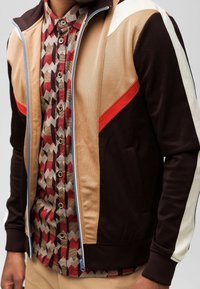 Vetoketjullinen urheiluvaunu ruskeana, beige- ja valkoisena punaisilla yksityiskohdilla; alla on geometristen kuvioiden t-paita ja sivutaskut.