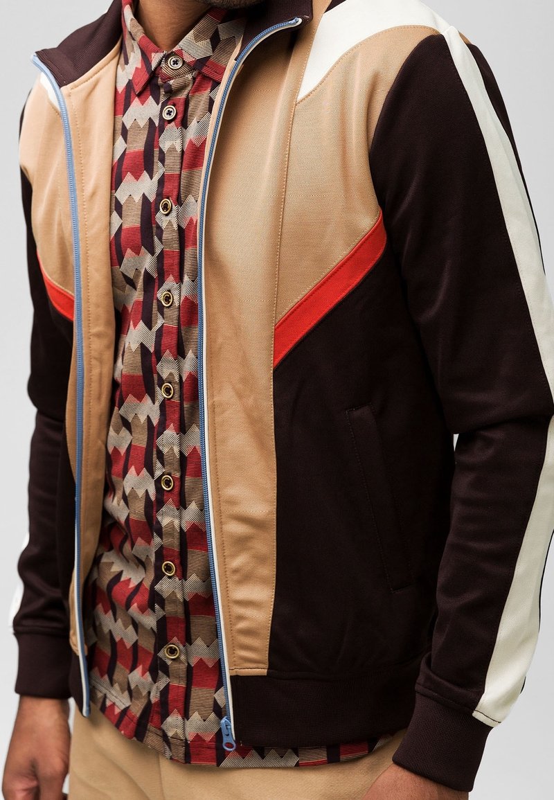 Vetoketjullinen urheiluvaunu ruskeana, beige- ja valkoisena punaisilla yksityiskohdilla; alla on geometristen kuvioiden t-paita ja sivutaskut.