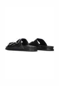 Zwarte leren instapsandalen met oversized decoratieve flappen en ronde accenten, vlakke zool, open achterkant en een gladde textuur.