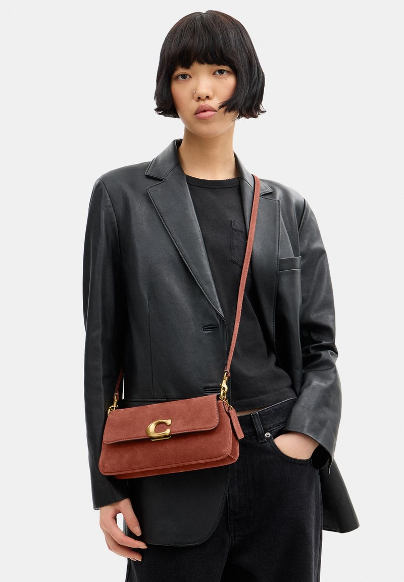 Femme aux cheveux courts noirs porte un blazer en cuir noir et un jean, portant un sac bandoulière en daim couleur rouille avec une fermeture dorée.