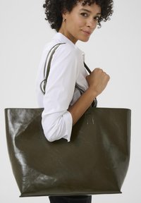 Sac fourre-tout en cuir vert olive avec une finition brillante, présentant deux poignées robustes et un détail de couture rectangulaire sur le côté.