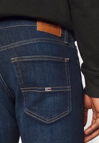 Bakfickan på mörkblå Tommy Jeans denim med synlig sömnad och liten broderad Tommy Hilfiger-flagga, bärs med svart tröja.