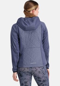 Veste softshell bleu marine avec un panneau matelassé texturé au dos, dotée d'une capuche et d'une coupe ajustée. Détails de coutures uniformes présents.