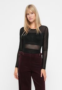 Donna bionda che indossa un top lungo a maniche nere trasparenti e pantaloni in velluto a coste viola scuro a vita alta, in piedi davanti a uno sfondo bianco.
