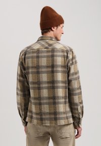 Cămașă din flanel pătat maro, cu guler, mâneci lungi butonate la manșete, realizată dintr-un material texturat și cald, purtată cu o caciulă tricotată maro.