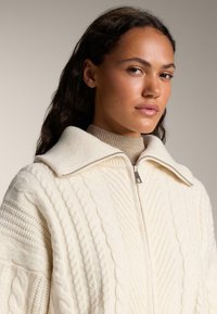 Pull crème en tricot torsadé avec un grand col, présentant une fermeture éclair sur toute la longueur et des motifs texturés sur l'ensemble du tissu. Fini lisse.