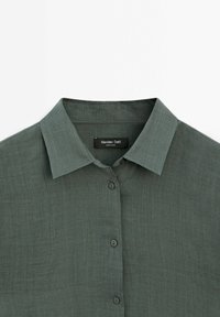 Zelená lněná košile s knoflíkovým zapínáním vpředu, klasickým límečkem a jemnou texturou. Štítek nese nápis "Massimo Dutti".