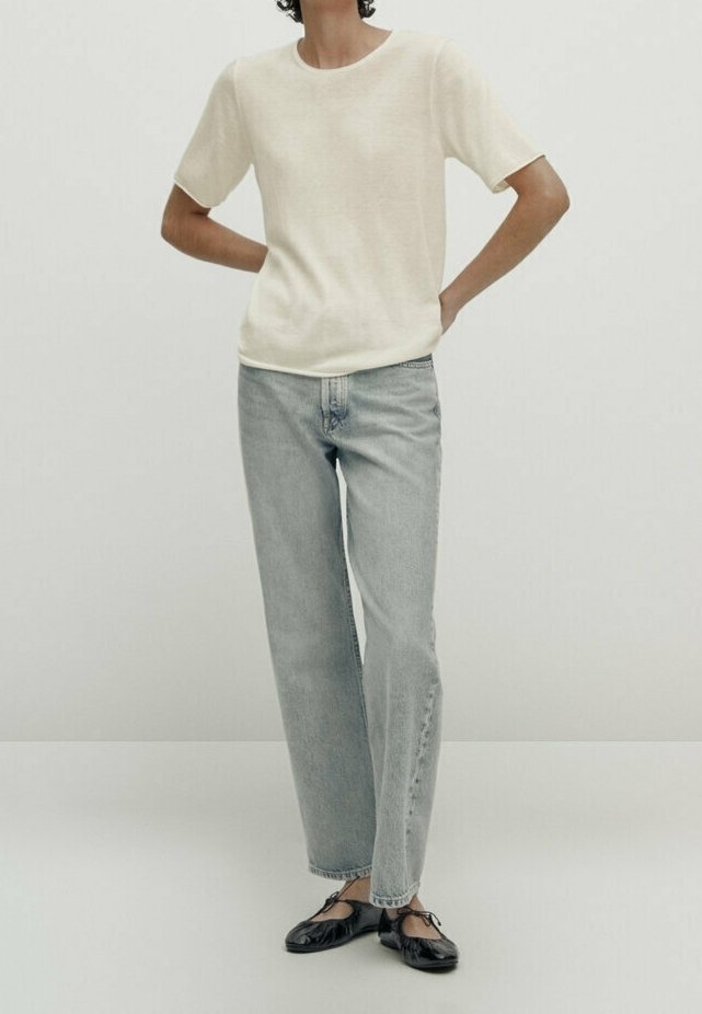 Personne portant une chemise beige à manches courtes, un jean bleu clair à jambes droites et des ballerines noires, debout les mains sur les hanches.