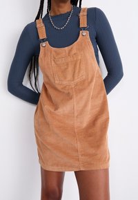 Latzkleid aus Cord in Hellbraun, mit Schulterträgern aus Metall, Fronttasche und einem strukturierten Rippmuster.