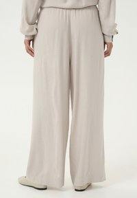 Pantalons large en beige clair avec une texture douce et fluide. Taille élastique et détail de couture subtil à l'ourlet. Associés à des chaussures beiges.