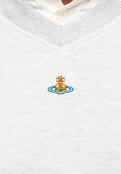Λευκό πουλόβερ με V-λαιμόκοψη και γιακαδάκι από πουκάμισο, με μικρό κέντημα χρυσής κορώνας περιτριγυρισμένης από μπλε κύκλο στο στήθος.