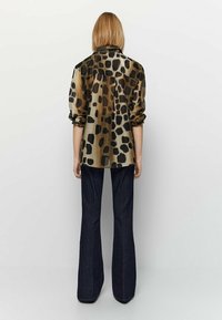 Koszula w panterkę z przezroczystego materiału, z podwiniętymi rękawami, w zestawie z ciemnymi spodniami w dzwony. Wzór posiada duże czarne plamy na brązowym tle.