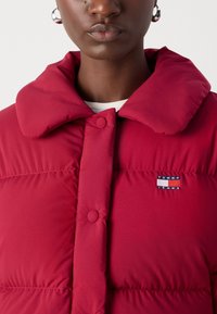 Κόκκινο φουσκωτό μπουφάν με μεγάλο γιακά, διαθέτει κουμπιά κατά μήκος του μπροστινού μέρους και μικρό λογότυπο "TOMMY JEANS" σε μπλε, λευκό και κόκκινο χρ�ώμα. Λείος υφάσ.