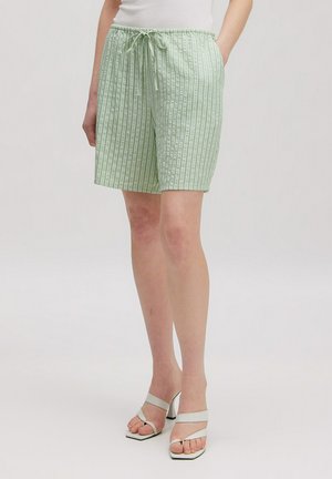 Persoon die lichtgroene gestreepte shorts met koord draagt en witte sandalen met hoge hakken, staand tegen een effen achtergrond.
