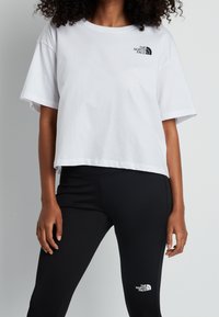 Vit kortärmad t-shirt med en logotyp på bröstet, kombinerad med svarta leggings som har samma logotyp på vaden.