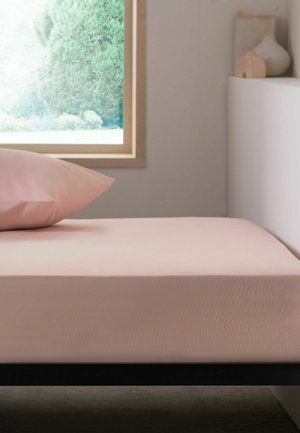 Einzelbett mit pinken Laken und Kissen neben einem Fenster mit Blick auf grüne Bäume, mit einem weißen Nachttisch, auf dem dekorative Vasen stehen.