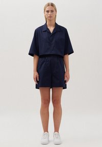 Marineblauwe korte mouwen blouse met kraag, gecombineerd met bijpassende shorts. Heeft een enkele borstzak, een ontspannen pasvorm en een zachte stoftextuur. Draag het met witte sneakers.