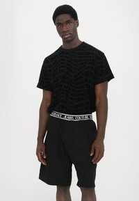 Juodas trumpas marškinėliai su viso paviršiaus Versace logotipo raštu, derinami su juodomis šortais. Juostos viršuje matyti užrašas "Versace Jeans Couture".