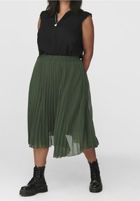 Svart ärmlös blus med volangkrage, kombinerad med en grön plisserad midikjol. Svarta snörstövlar fulländar outfiten.