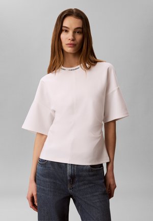Giovane donna con capelli castani lisci che indossa una maglietta bianca oversize a maniche corte con il logo Calvin Klein sul colletto e jeans blu.