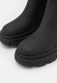 Zwarte rubberen enkellaarzen met een matte afwerking, een ronde neus, een dikke contrasterende zool en een gestructureerd profiel.