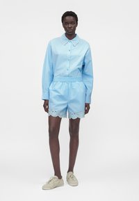 Femme portant une chemise à manches longues bleu clair avec un col festonné et un short assorti avec bordure en dentelle ajourée, chaussures beige à lacets, debout sur fond blanc.