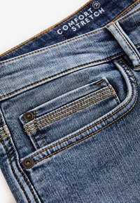 Detailný záber na modrú džínovú kapsu s oranžovým prešitím a štítkom "Comfort Stretch" vo vnútri pása.