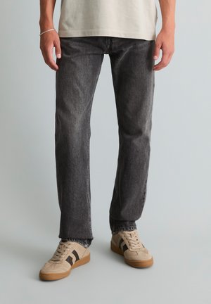 Mann trägt ausgewaschene schwarze Straight-Leg-Jeans, beige Sneakers mit schwarzen Streifen und ein beiges Hemd, steht vor einem einfarbigen Hintergrund.
