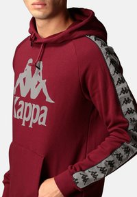 Felpa bordeaux con un grande logo Kappa grigio sul davanti, caratterizzata da strisce grigie di accento con un motivo del logo ripetuto lungo le maniche.