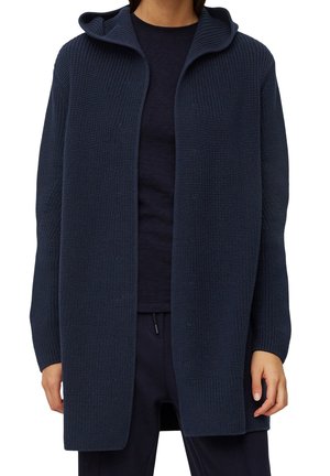 Persoon draagt een marineblauwe, opengebreide cardigan met capuchon, over een bijpassend donkerblauw shirt en broek.