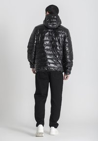 Chaqueta acolchada negra con un acabado brillante y capucha, que presenta secciones acolchadas. Combinada con pantalones negros y zapatillas blancas, mostrada desde la parte posterior.