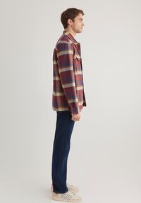 Kariertes Hemd in Rottönen, Blau und Beige, mit zwei Brusttaschen, kombiniert mit dunkelblauen Jeans und neutralen Sneakers.