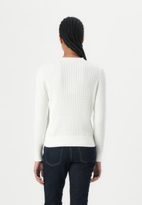 Hvit strikkegenser med et strukturert mønster, ribbestrikkede ermer og nedkant. Kombinert med mørke denimjeans, sett fra baksiden.
