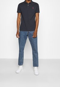 Polo shirt marine avec un logo rouge, manches courtes et col, associé à un jean bleu et des baskets blanches. Design d'une tenue décontractée.