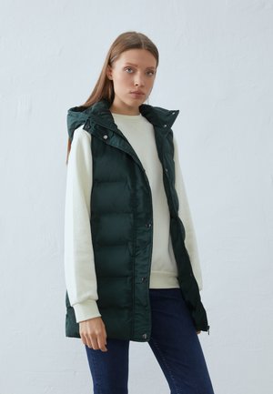 Nabity zielony kamizelka z kapturem, z przodu zapinana na zamek błyskawiczny i napy, noszona na kremowym swetrze. Gładka tekstura, długość do kolan.