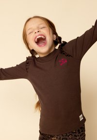 Bruine geribde top met lange mouwen, voorzien van ruffles op de schouders en een roze geborduurd bericht, gecombineerd met een patronenlegging.