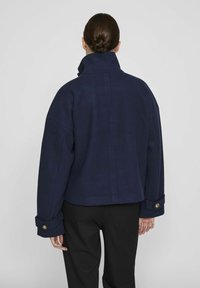 Mørkeblå jakke med høy krage, brede ermer og knappede mansjetter. Den myke teksturen og den forkortede designen fremhever den moderne silhuetten.