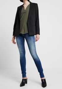 Sort dobbeltradet blazer over olivengrøn bluse, parret med mellem-høj talje blå jeans og sorte åbne hæle.