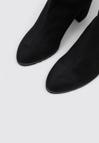 Botines de ante negros con punta afilada y tacón ancho. Presentan una textura suave y costuras verticales en la parte delantera.
