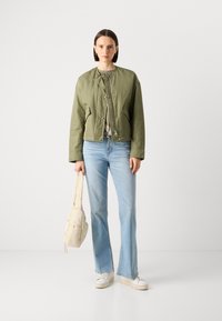Giubbino bomber corto verde oliva con collo arricciato, abbinato a jeans svasati azzurri chiari e una borsa beige. Sneakers bianche completano il look.