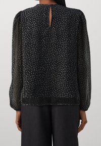 Svart blus med en texturerad, mönstrad design med ljusgrå prickar, långa ärmar och ett nyckelhål bak. Mjuk, genomskinlig tyg.